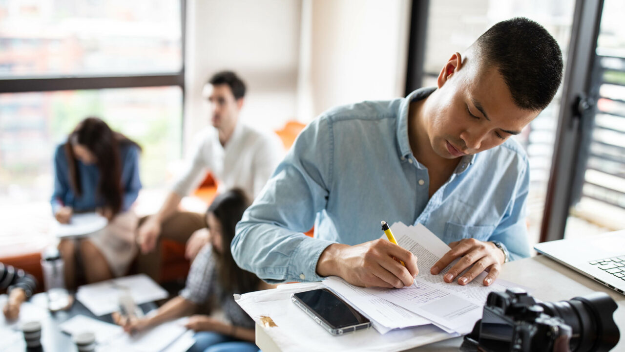 Photo of a man in an office. He is making notes on a document with a pencil and there are three other people in the background having a conversation.