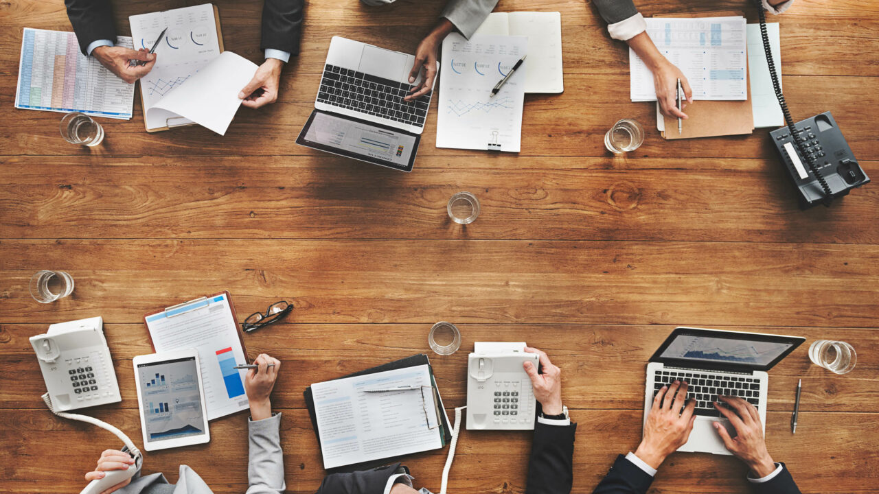 Photo of six people sitting at a conference room table using laptops and reviewing paperwork