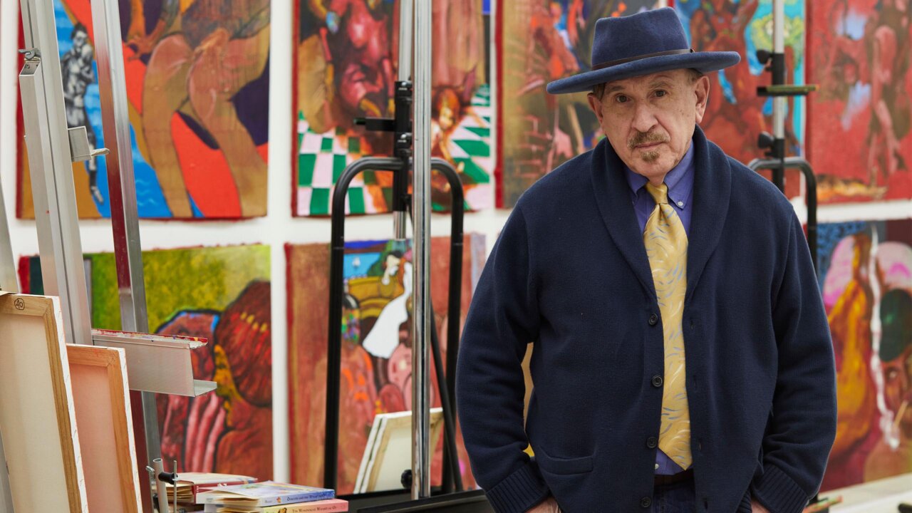 Archie Rand standing in front of an exhibition of his paintings (Photo Credit: Kevin Blumenthal)